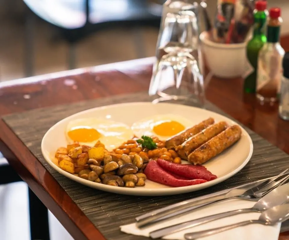 Cooked breakfast on a white plate: two fried eggs, three grilled sausages, slices of beef salami, sautéed mushrooms, diced roast potatoes and baked beans garnished with parsley, with cutlery on a napkin and upturned glasses on a polished wooden table