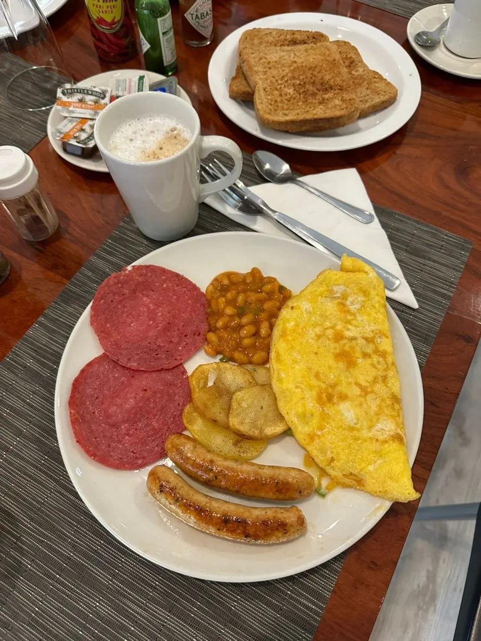 Plated full breakfast of beef salami, baked beans, fried potato slices, a folded omelette and two grilled sausages, served with a cappuccino and a plate of brown toast on a wooden table
