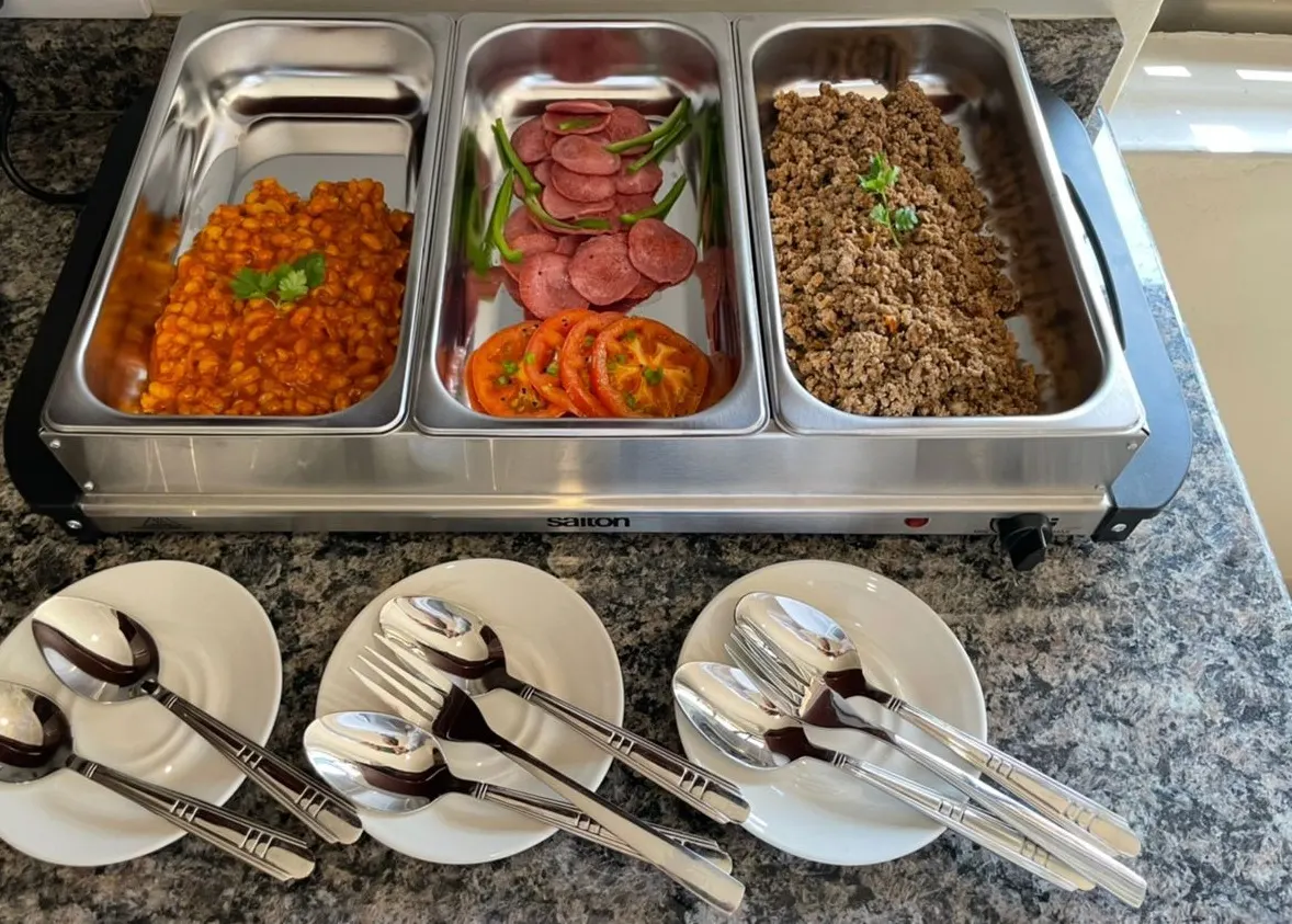 Hot breakfast buffet warmer on a granite counter with three steel trays: baked beans garnished with fresh herbs, sliced beef salami with tomato slices and green beans, and savoury mince, with side plates of serving spoons and forks laid out in front