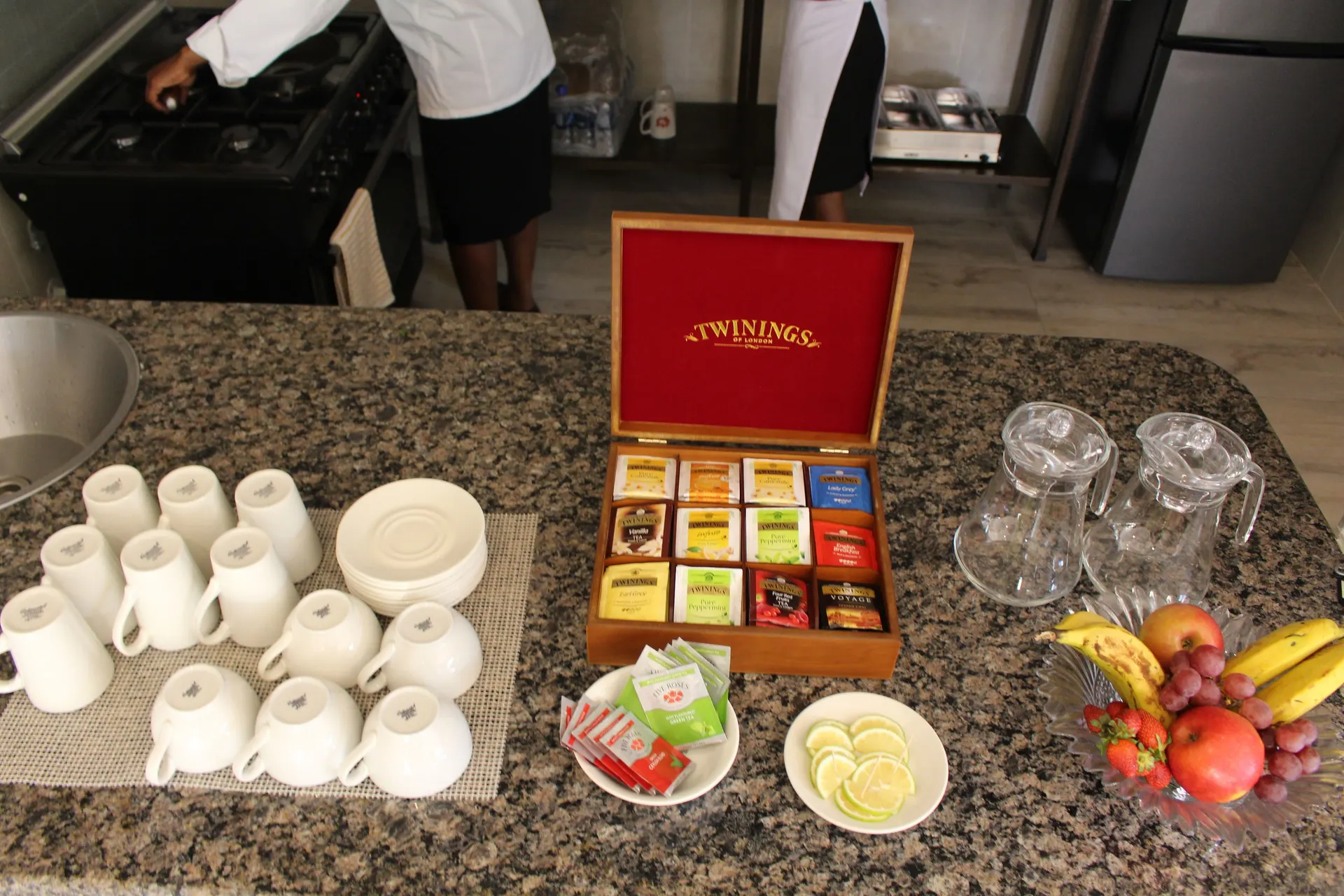 Tea station on a granite counter with an open Twinings tea box, rows of white cups and saucers, a plate of lemon slices, tea sachets, empty glass jugs and a bowl of apples, bananas, grapes and strawberries