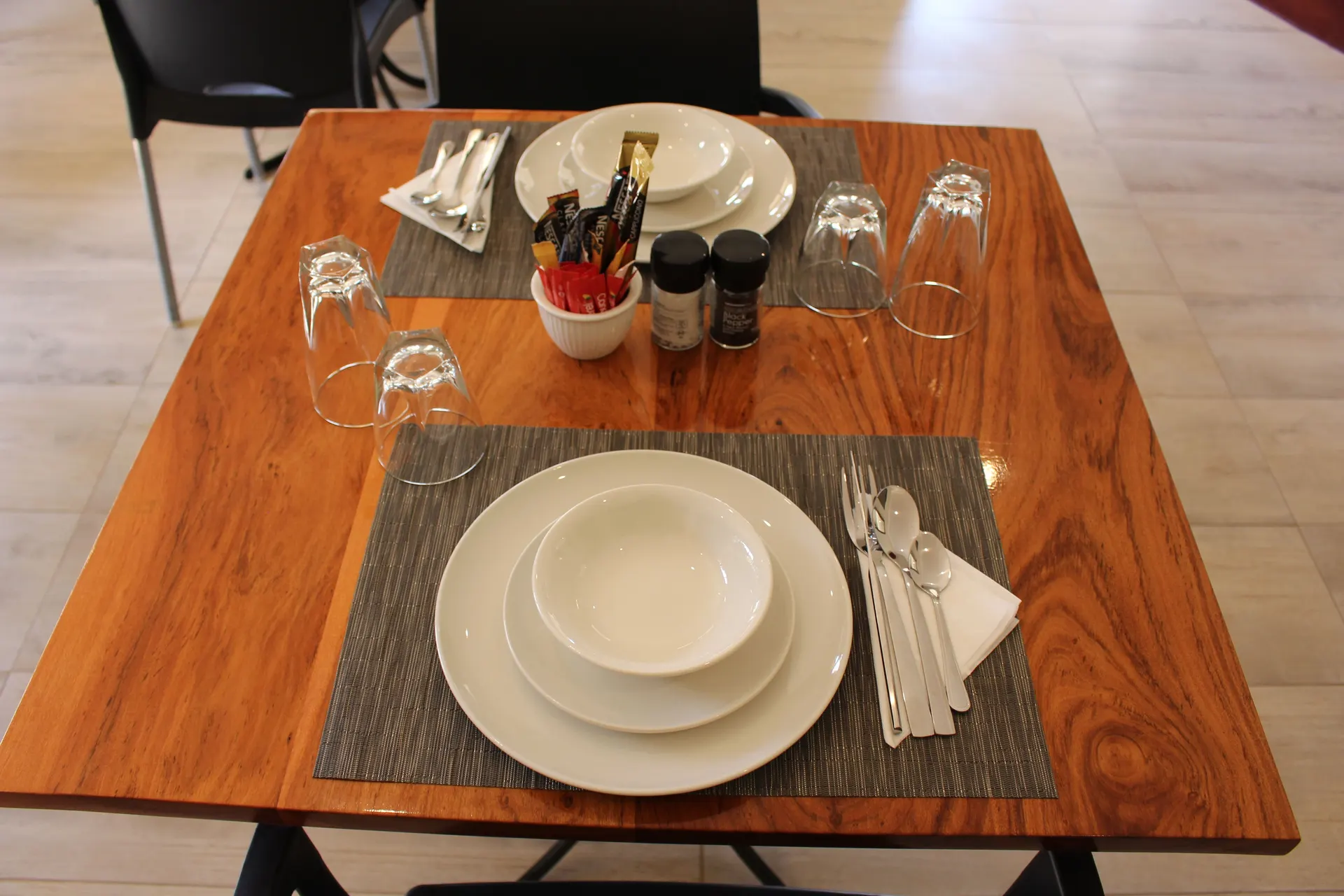 Overhead view of a table for two laid for breakfast, with white plates and bowls on grey woven placemats, cutlery on napkins, upturned glasses, salt and pepper grinders and sugar sachets on a warm wood-grain table