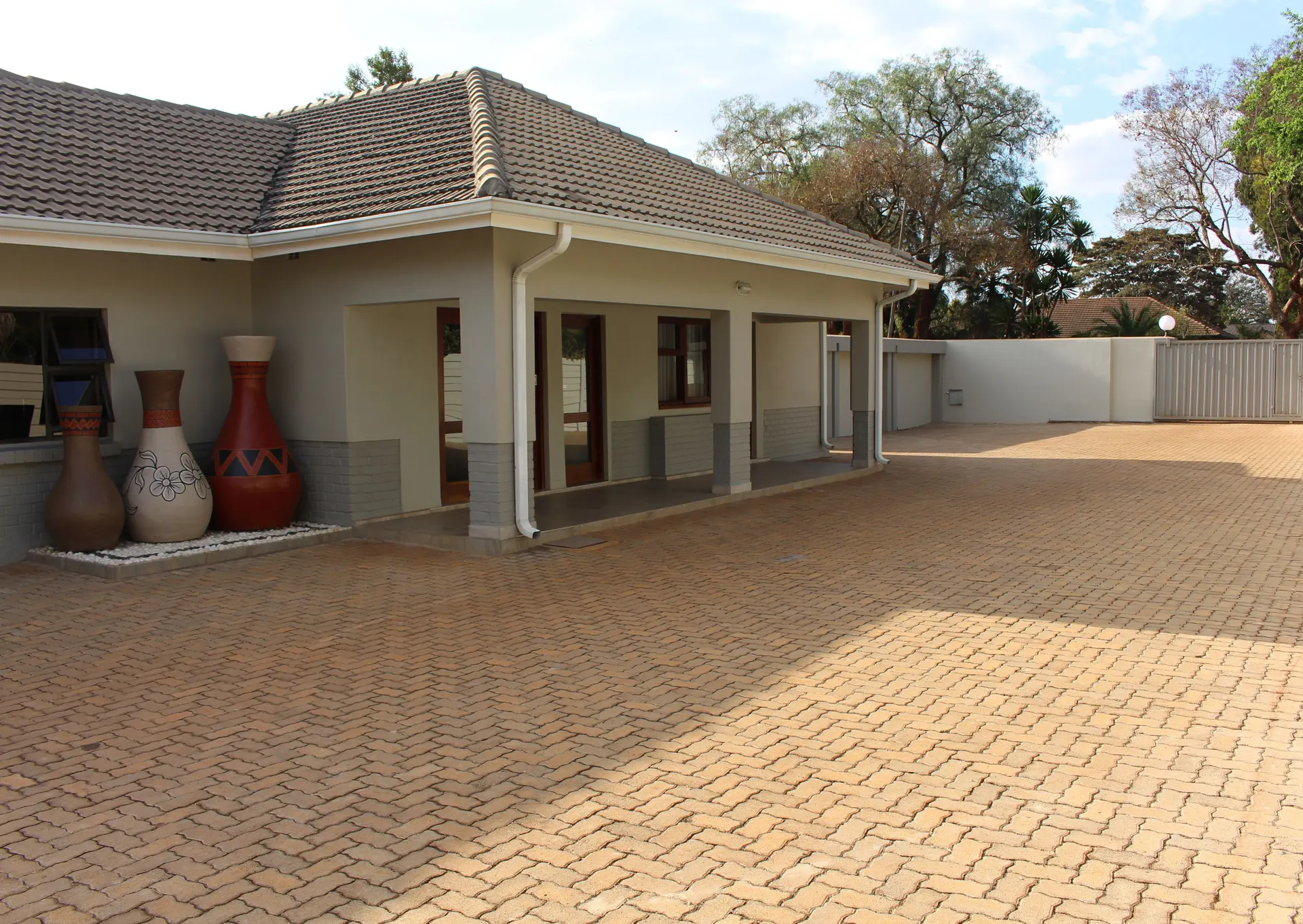 Covered veranda of a FIRN Lodge guest wing with three decorative African urns beside the paved courtyard in late-afternoon sun, with the entrance gate beyond