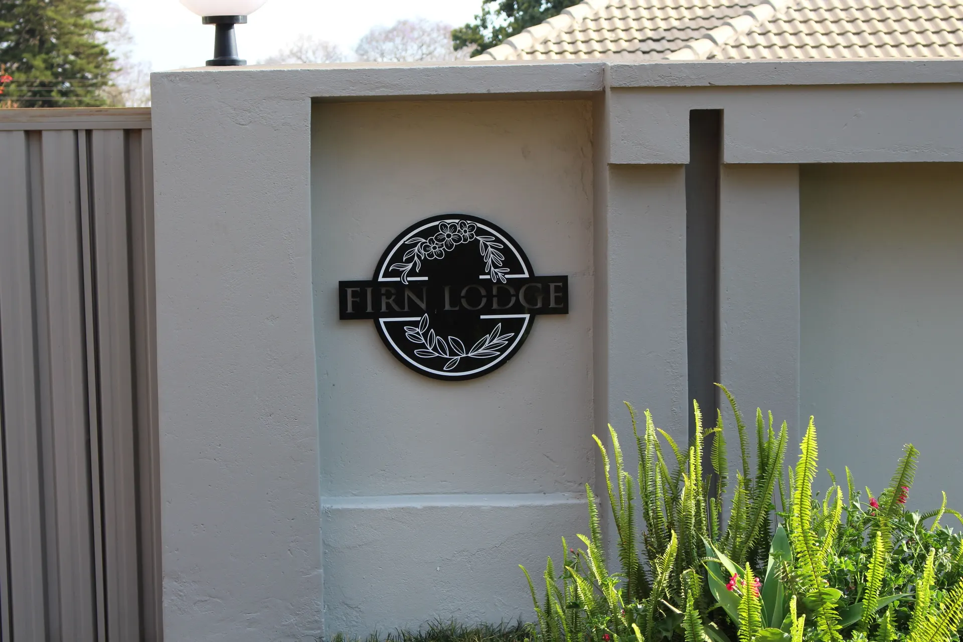 Round black FIRN Lodge sign with a floral wreath on the grey gate pillar, framed by green ferns