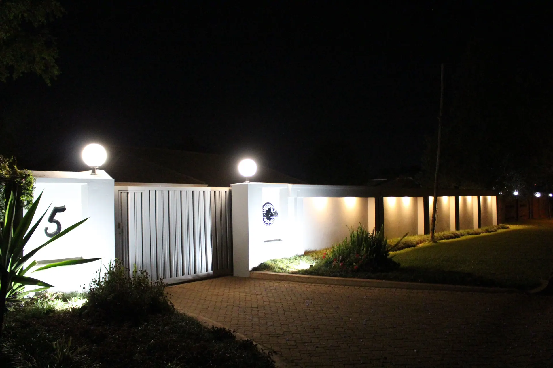 FIRN Lodge entrance at night: glowing globe lamps on the gate pillars, house number 5, the round lodge sign and a row of warm lights washing the boundary wall