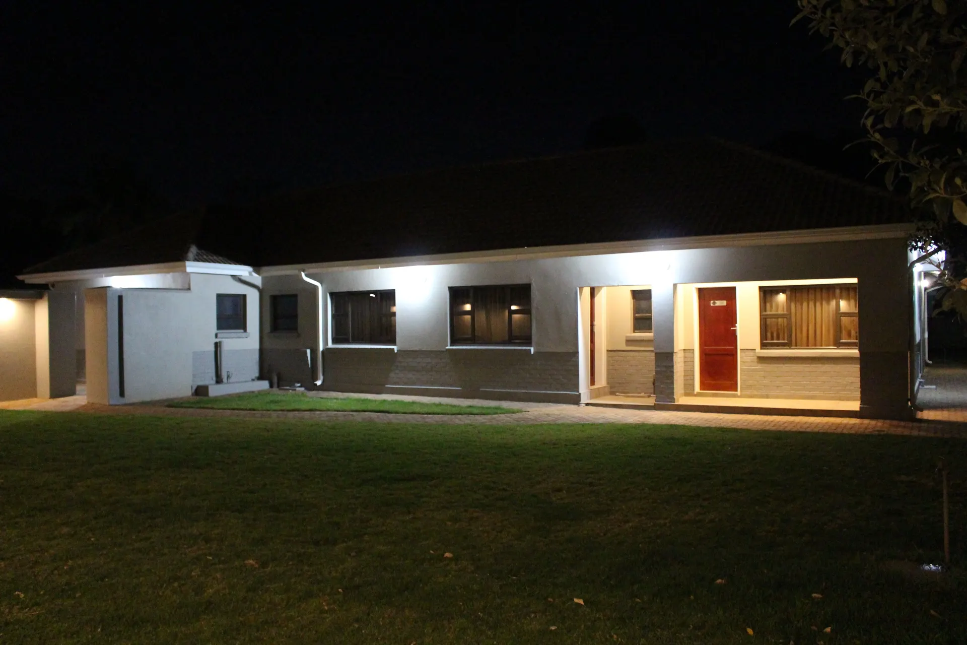 FIRN Lodge rear guest wing at night seen across the lawn, with a lit veranda, a red room door and warmly glowing curtained windows