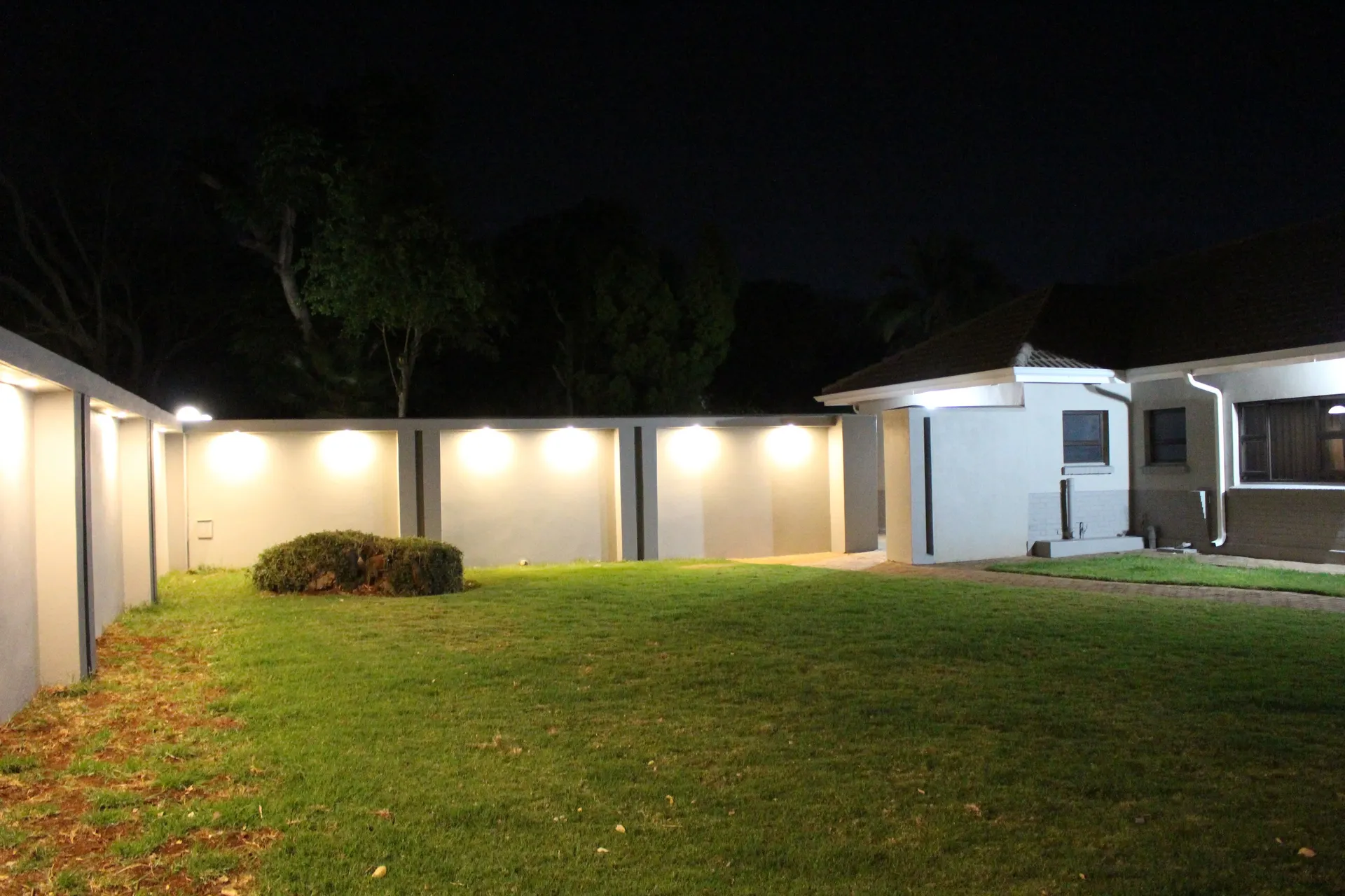 FIRN Lodge garden at night: a lawn and rounded shrub enclosed by a garden wall washed with warm lights, with the corner of a guest wing on the right