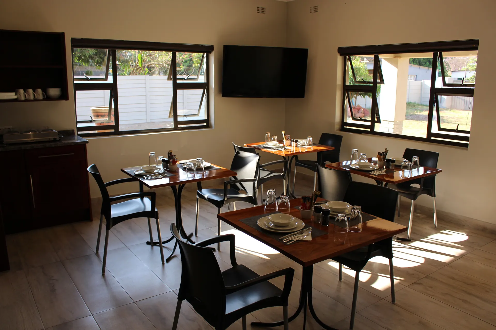 Sunlit breakfast room with four polished timber tables laid for breakfast, black chairs, a wall-mounted TV between two windows looking out to the garden, and a kitchenette counter with a crockery shelf on the left